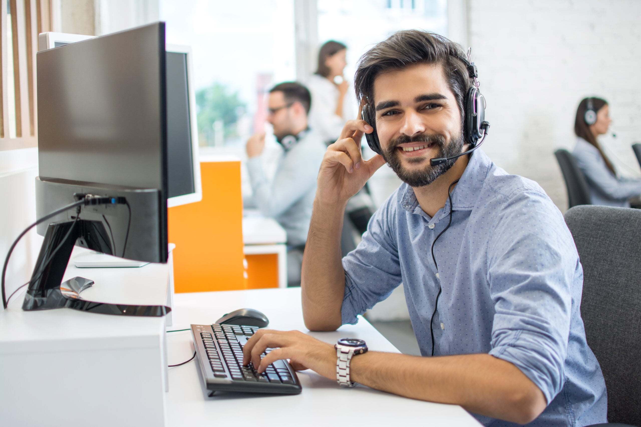 a beard confident professional customer service representative wearing a headphone in front of his computer with some colleague at the back ground working at the office