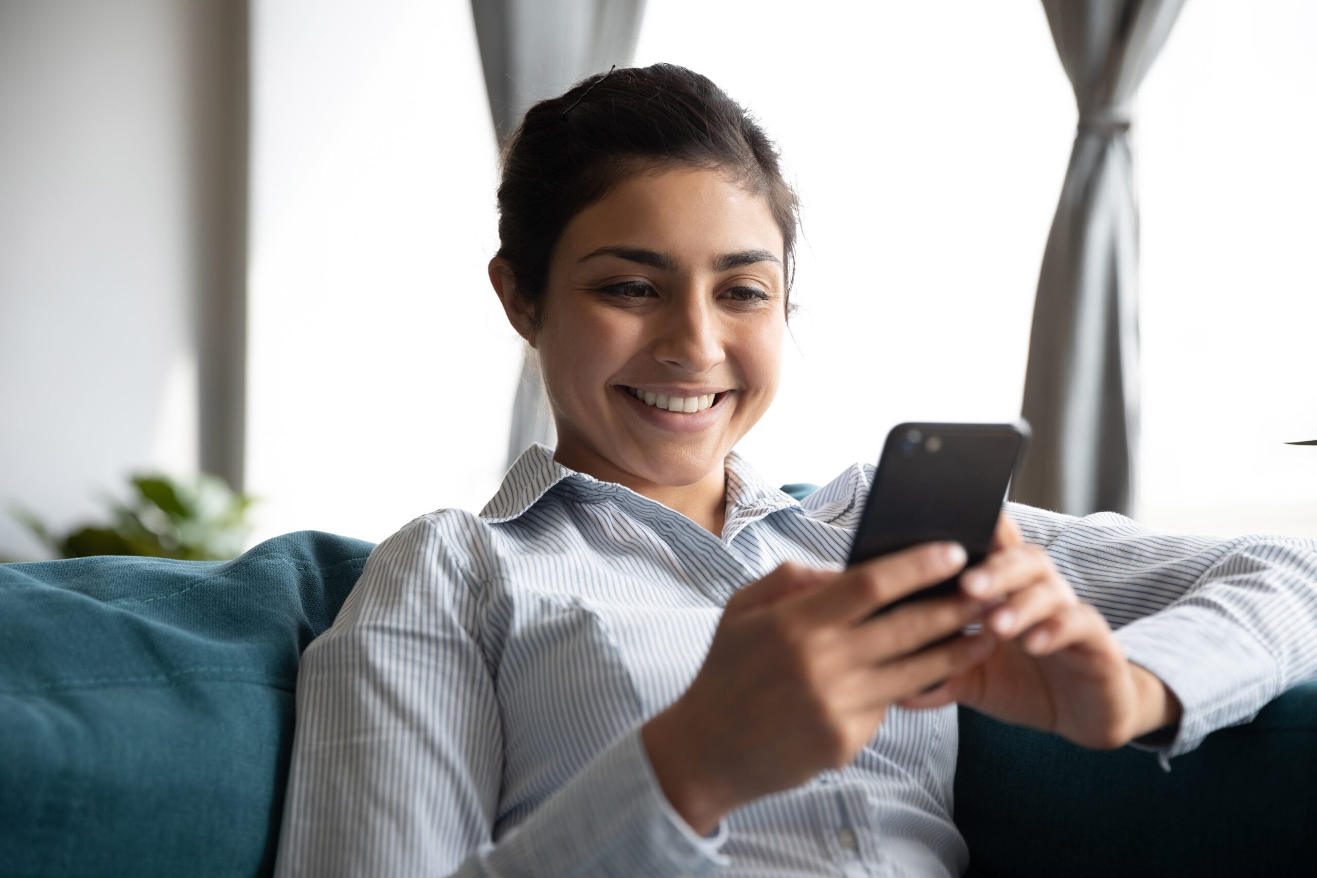a smiling woman sitting in a blue sofa while using her mobile phone