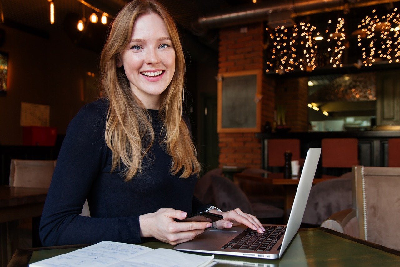 a delighted women sitting in a chair holding a mobile phone and her laptop on the table with some lights at the background
