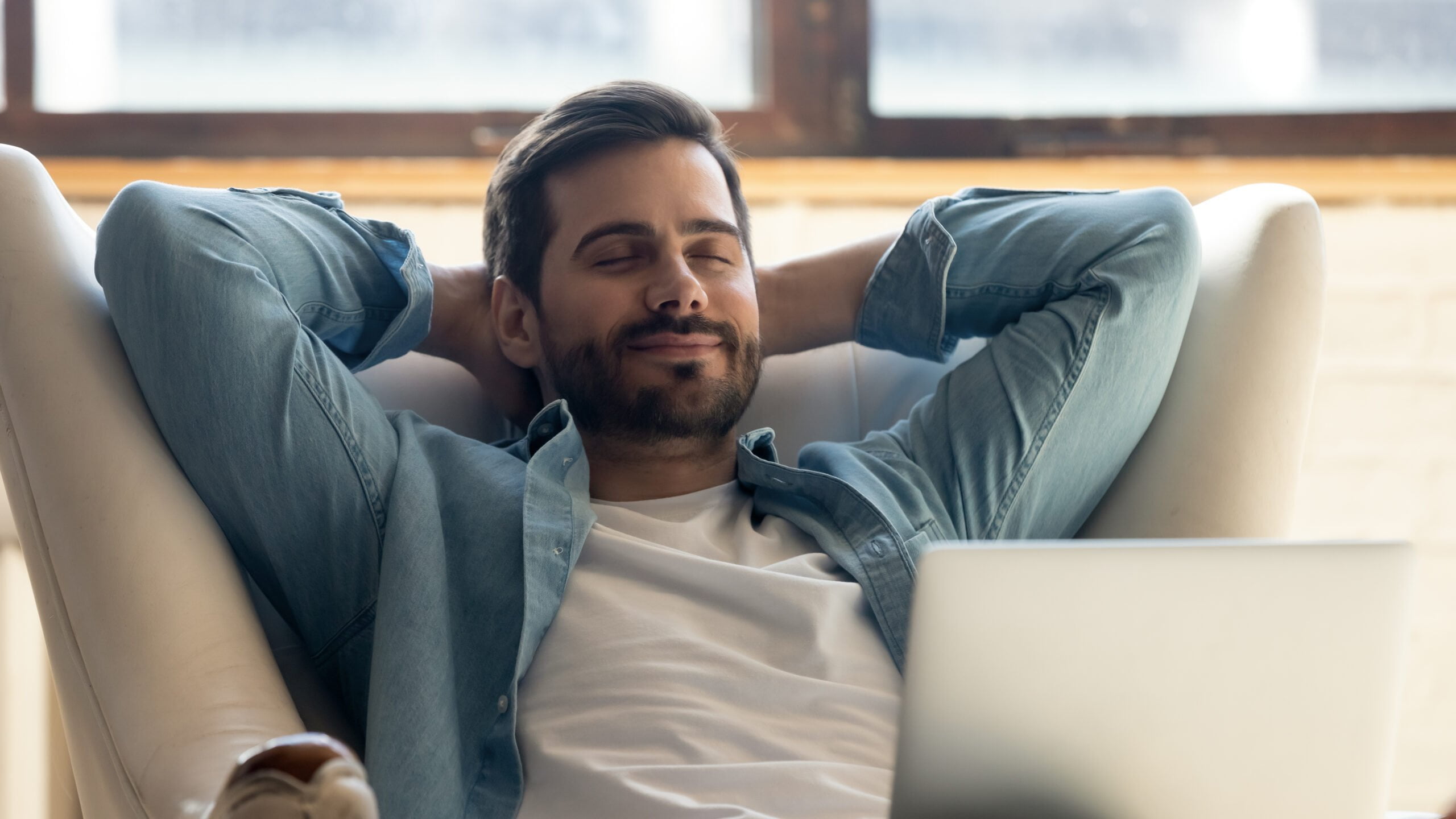 a beard relaxed man sitting in a white armchair wearing a blue long sleeve shirt