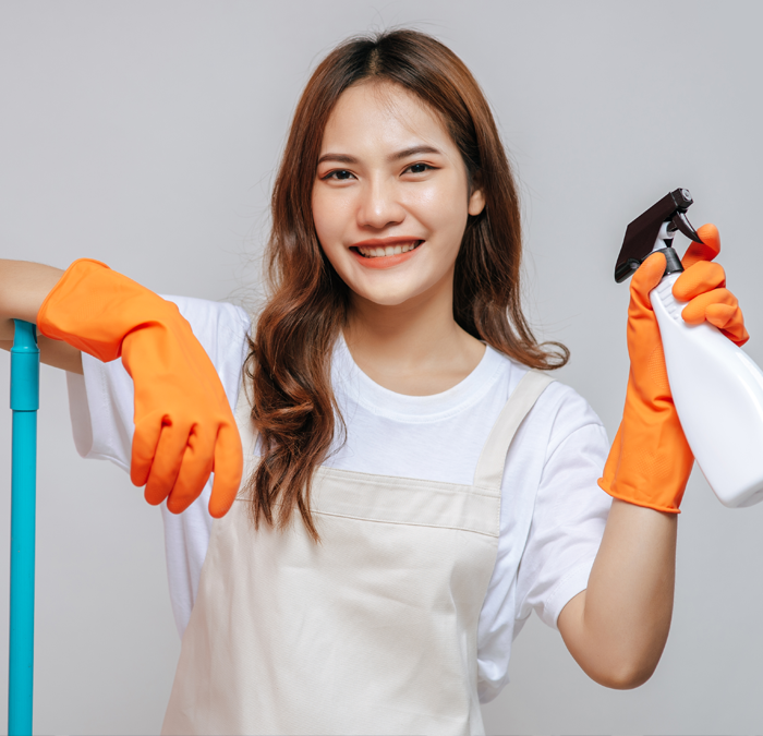 a smiling professional cleaner wearing an apron over a white t-shirt holding a spray cleaning bottle with her orange gloves